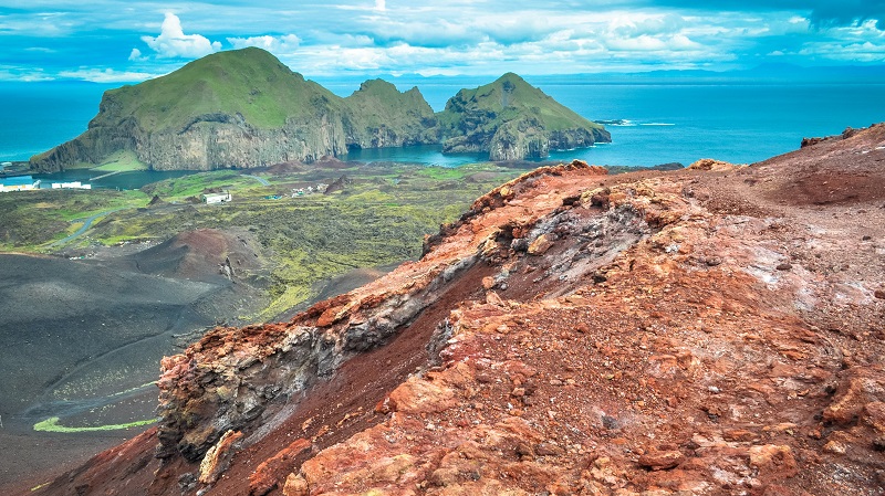Vulkaniskt landskap på Heimaey, med rödaktig berggrund och några gräsgröna småöar precis utanför.